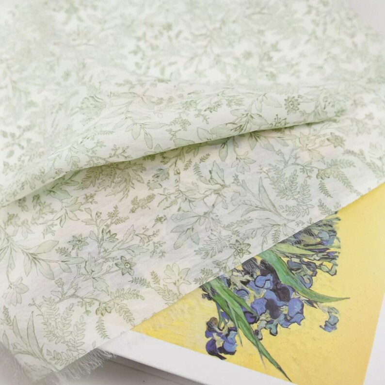Puede incluir: Una tela floral verde claro y blanca con un delicado estampado de hojas. La tela est&aacute; drapeada sobre una impresi&oacute;n de la pintura "Lirios" de Van Gogh, con lirios azules sobre un fondo amarillo.