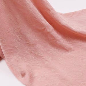 May include: A soft, dusty pink fabric featuring a subtle, tone-on-tone jacquard floral pattern. The material displays a delicate, crinkled texture with gentle folds and drapes, creating a soft play of light and shadow across the surface.