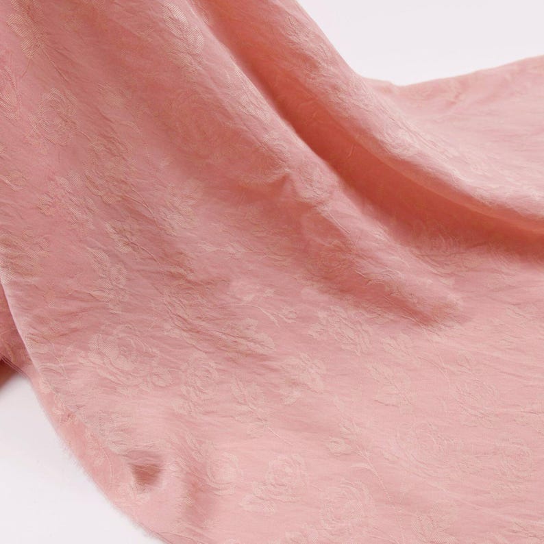 May include: A soft, dusty pink fabric featuring a subtle, tone-on-tone jacquard floral pattern. The material displays a delicate, crinkled texture with gentle folds and drapes, creating a soft play of light and shadow across the surface.