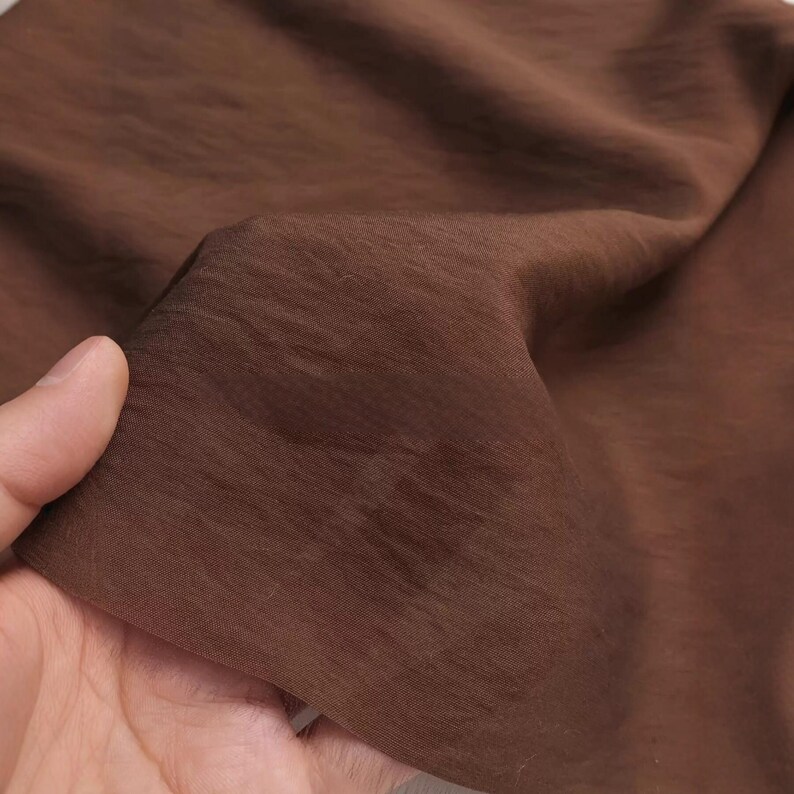 K&ouml;nnte beinhalten: Ein St&uuml;ck einfarbiger brauner Stoff mit einer sichtbaren, feink&ouml;rnigen Textur. Das Material weist subtile horizontale Falten und Knitter auf der Oberfl&auml;che auf, die das matte Finish und die gleichm&auml;&szlig;ige tiefbraune Farbe des Textils betonen.