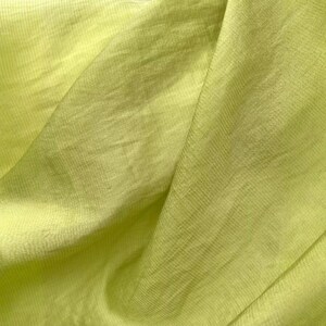 May include: A piece of light lime green fabric with a visible woven texture. The material is draped to create soft folds and shadows, highlighting the consistent color and the fine, interlaced pattern of the threads throughout the textile surface.