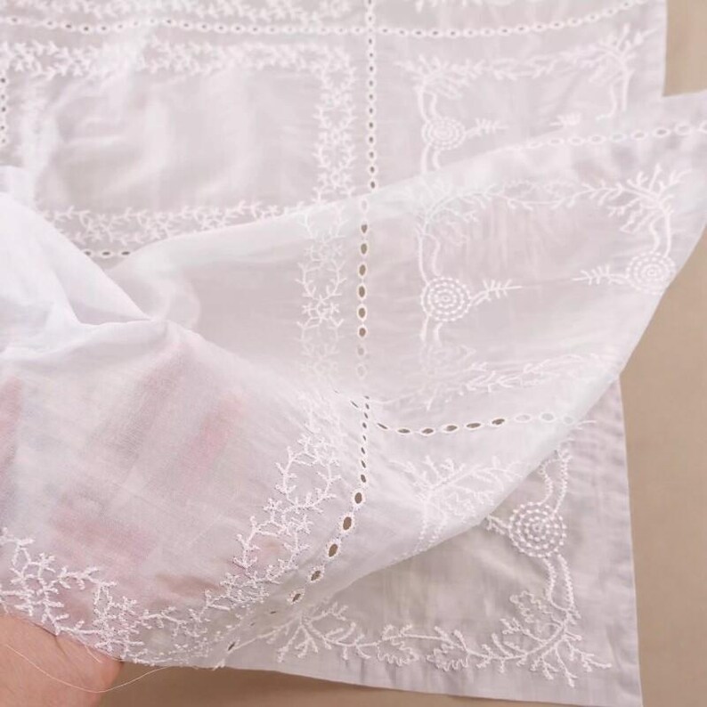 Op de afbeelding: Close-up van witte stof met ingewikkelde geborduurde bloemen- en geometrische patronen. De delicate borduursels cre&euml;ren een textuureffect, met een transparante kwaliteit voor de stof. Het ontwerp bevat bloemmotieven en geometrische vormen.
