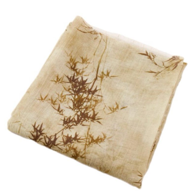 May include: A folded beige scarf with a brown bamboo leaf pattern. The scarf is made of a lightweight fabric and has a natural, organic aesthetic. The bamboo design is detailed and adds a touch of nature-inspired elegance.