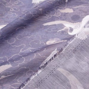May include: Close-up of a fabric with a repeating pattern of white cranes and clouds on a muted blue background. The fabric has a soft, flowing appearance, with visible texture and a raw edge.