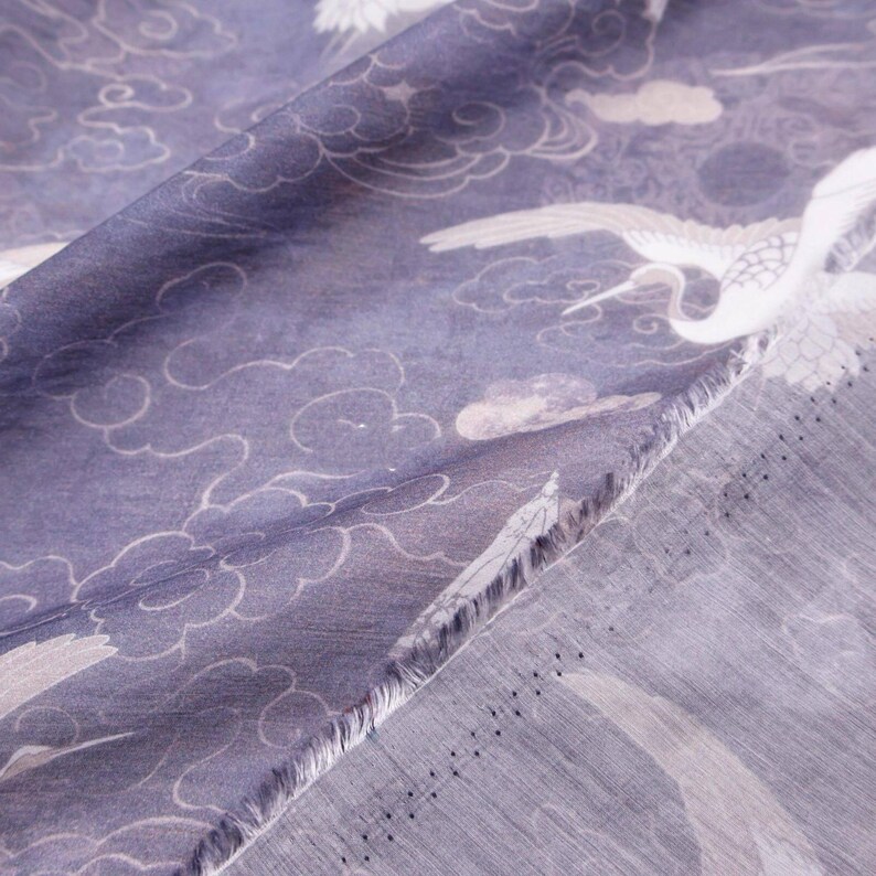 May include: Close-up of a fabric with a repeating pattern of white cranes and clouds on a muted blue background. The fabric has a soft, flowing appearance, with visible texture and a raw edge.