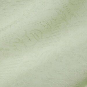 May include: Close-up of a pale green fabric with a subtle floral pattern. The fabric has a soft, textured appearance with visible folds and creases, suggesting it is a lightweight material. The design features delicate, repeating floral motifs.