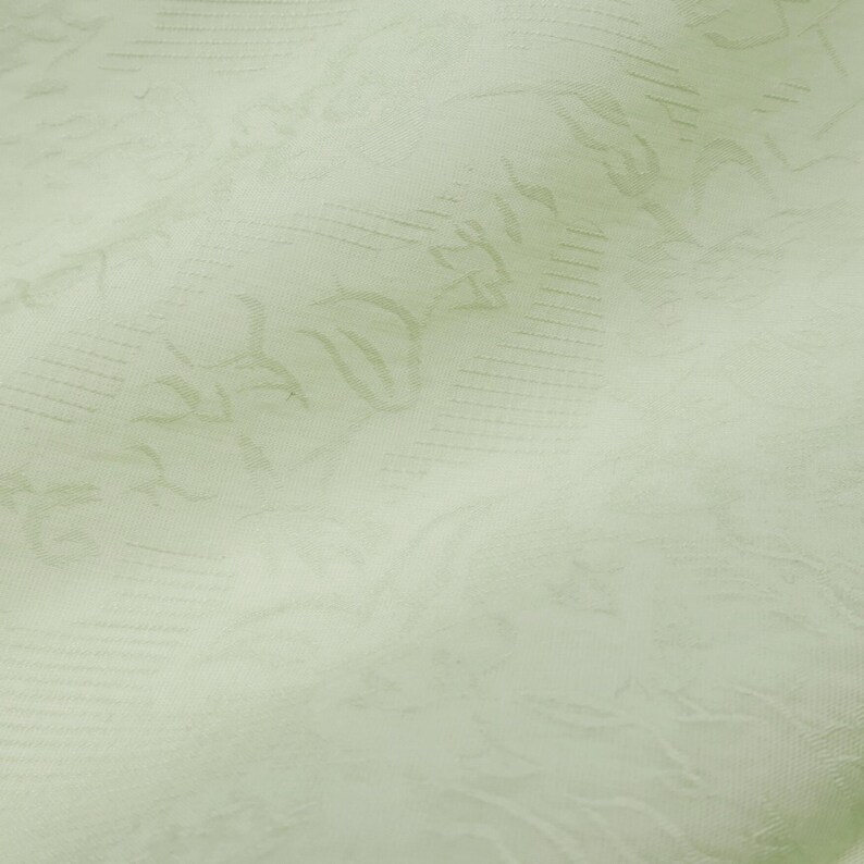 May include: Close-up of a pale green fabric with a subtle floral pattern. The fabric has a soft, textured appearance with visible folds and creases, suggesting it is a lightweight material. The design features delicate, repeating floral motifs.