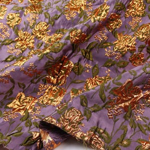May include: Close-up of a purple fabric with a floral pattern. The design features gold metallic roses and green leaves. The fabric has a textured appearance, with the metallic elements adding a shimmering effect. The overall aesthetic is luxurious and decorative.