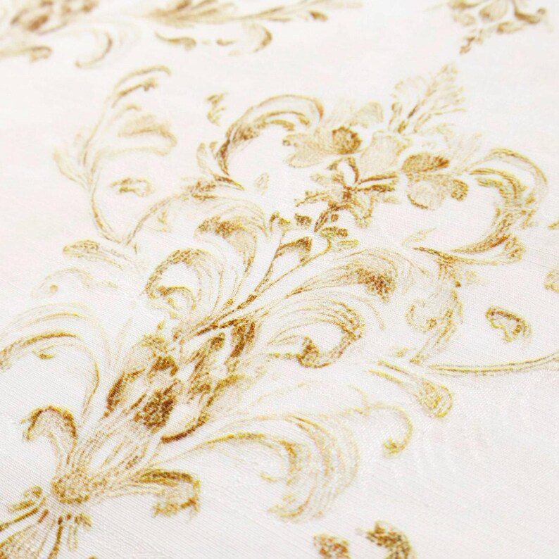 May include: Close-up of a white fabric with a repeating gold damask pattern. The design features ornate, swirling floral and leaf motifs, creating a textured, elegant appearance. The fabric has a subtle sheen, enhancing the gold details.