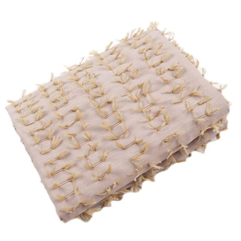 May include: A folded light beige textile throw blanket featuring a repeating pattern of small, vertical, tan-colored tufted fringe accents arranged in horizontal rows across the entire surface of the fabric, creating a textured appearance.
