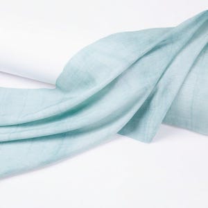 K&ouml;nnte beinhalten: Hellblauer Schal mit weicher Textur, drapiert auf wei&szlig;em Hintergrund. Der Stoff ist gefaltet und geschichtet, wodurch ein Gef&uuml;hl von Bewegung entsteht. Die Farbe ist ein sanftes, ged&auml;mpftes Blau.