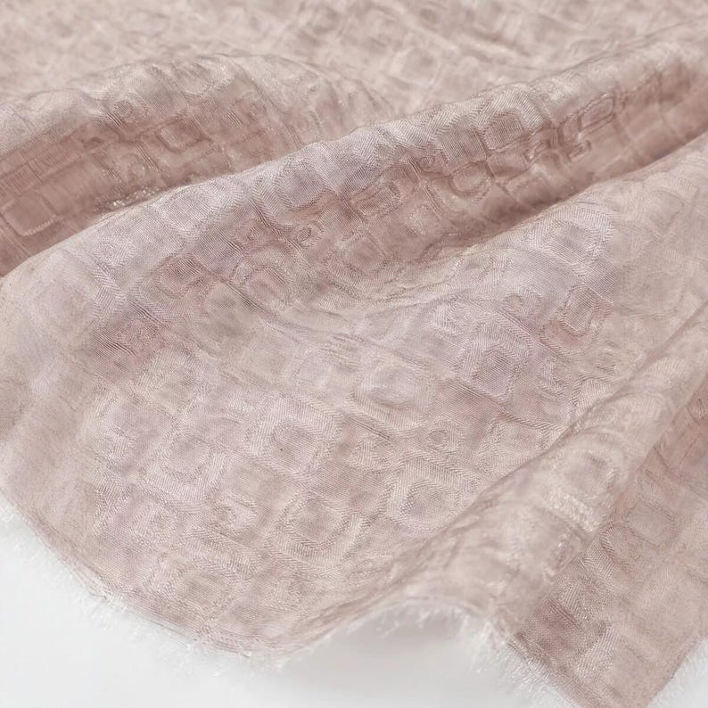May include: A close-up shot of a soft, flowing scarf in a muted pink hue. The fabric has a subtle, textured pattern of small squares, creating a delicate, almost ethereal appearance. The edges are slightly frayed, adding to the scarf's gentle aesthetic.