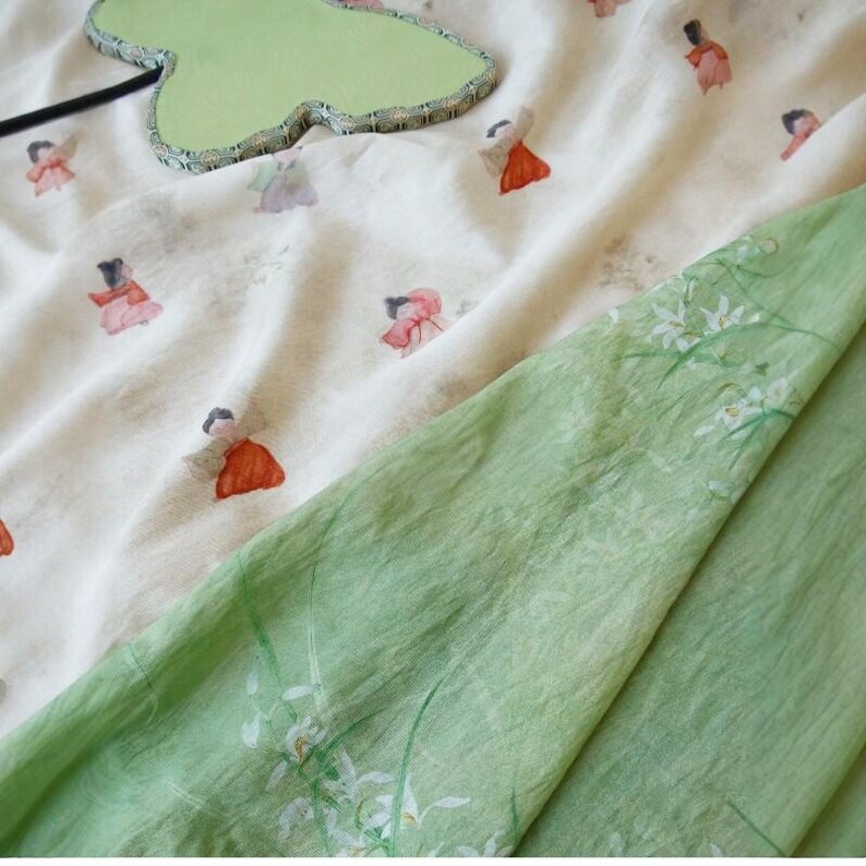 Peut inclure: Un tissu de couleur claire avec un motif r&eacute;p&eacute;t&eacute; de petites figures en robes rouges et roses. Un tissu vert avec un motif floral est drap&eacute; sur le tissu &agrave; motifs. Un objet vert en forme de feuille est dans le coin sup&eacute;rieur gauche.