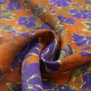 May include: Close-up of a fabric with a floral pattern. The fabric has a burnt orange background with large purple flowers and green and brown leaves. The fabric is folded, showing the texture and detail of the design.