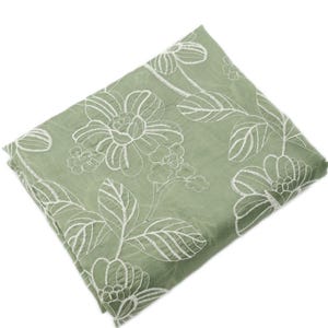 May include: A sage green fabric square featuring white embroidered floral patterns. The design consists of large, stylized blossoms and leafy stems stitched in a continuous white thread, creating a textured contrast against the solid green base.