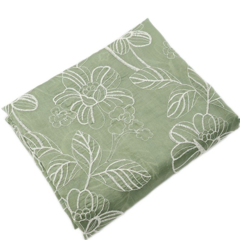 May include: A sage green fabric square featuring white embroidered floral patterns. The design consists of large, stylized blossoms and leafy stems stitched in a continuous white thread, creating a textured contrast against the solid green base.