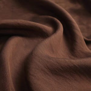 K&ouml;nnte beinhalten: Ein einfarbiger, dunkelbrauner Stoff mit einer sichtbaren Webstruktur, der durch weiche Falten und tiefe Schatten eine Reihe von wellenf&ouml;rmigen Kurven auf der Oberfl&auml;che bildet und das matte Finish sowie den tiefen Erdton betont.