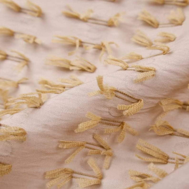 May include: A light beige fabric featuring a textured surface with evenly spaced, small, pale yellow tufted fringe accents. The material has a soft, slightly wrinkled appearance with the tufts arranged in a repeating pattern across the textile.