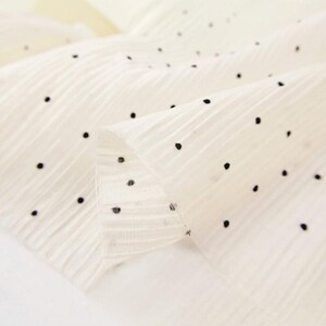 May include: Close-up of a cream-colored fabric with a delicate texture and small, evenly spaced black polka dots. The fabric is folded, revealing its layered construction and subtle sheen. Ideal for apparel or craft projects.
