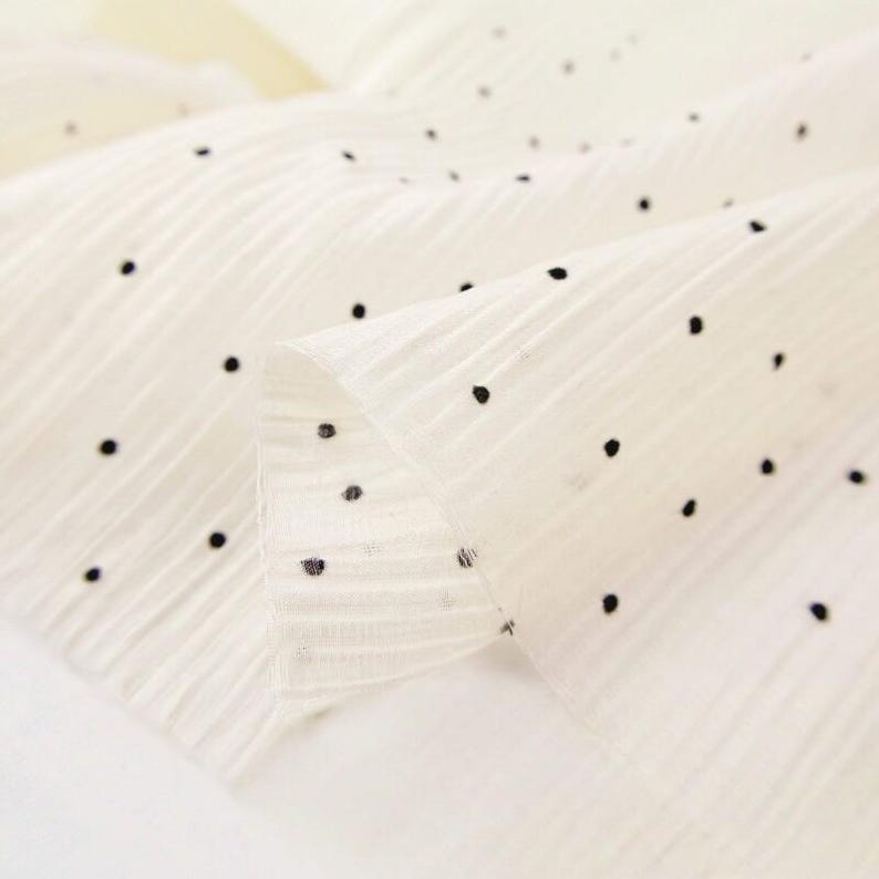 May include: Close-up of a cream-colored fabric with a delicate texture and small, evenly spaced black polka dots. The fabric is folded, revealing its layered construction and subtle sheen. Ideal for apparel or craft projects.