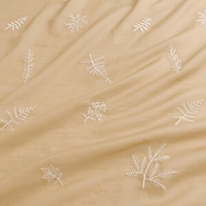 May include: A close-up of a tan fabric with white embroidered floral and leaf patterns. The fabric is slightly wrinkled, creating shadows and depth. The embroidery is detailed, with various botanical designs scattered across the surface.