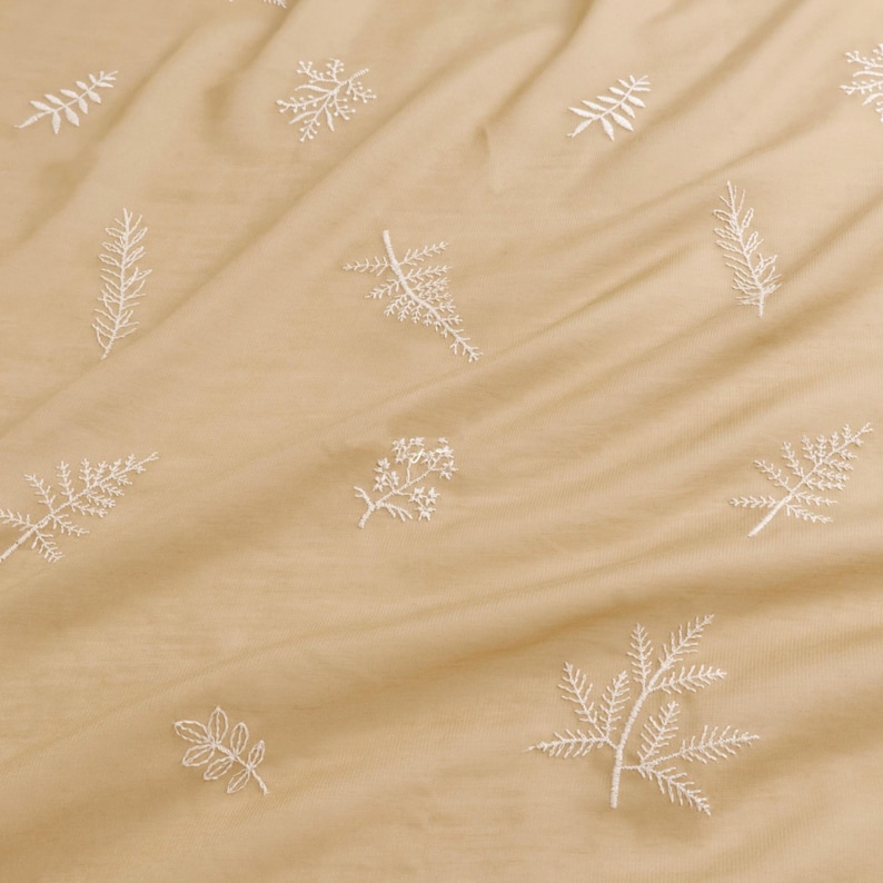 May include: A close-up of a tan fabric with white embroidered floral and leaf patterns. The fabric is slightly wrinkled, creating shadows and depth. The embroidery is detailed, with various botanical designs scattered across the surface.