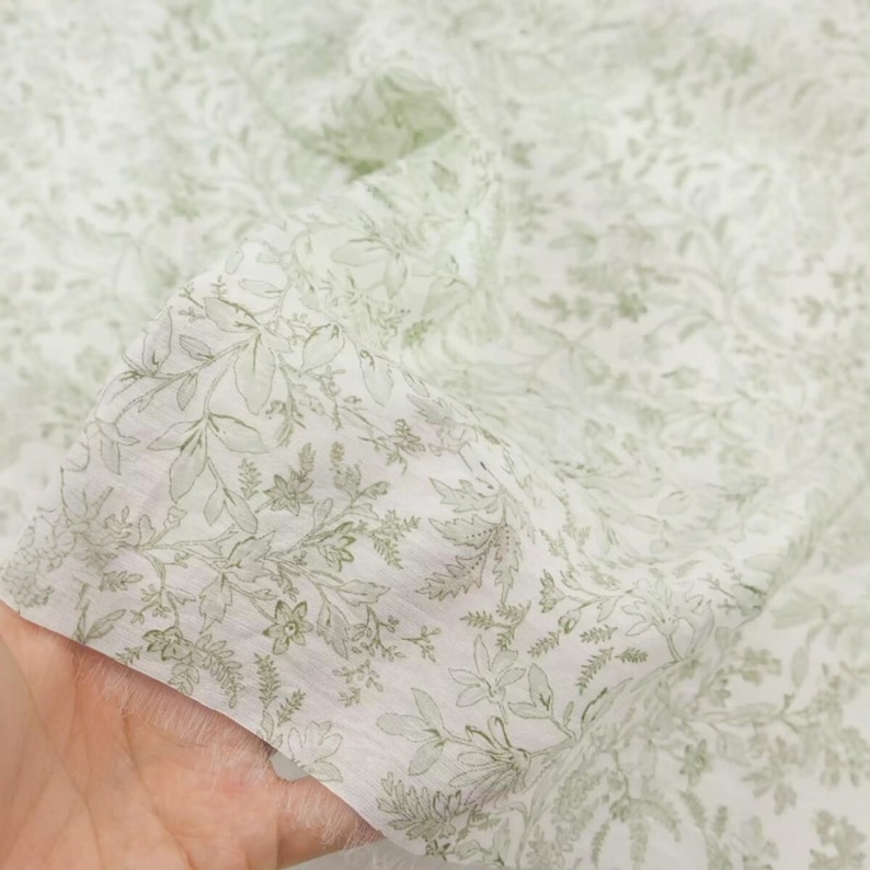 Puede incluir: Primer plano de una tela blanca con un delicado estampado floral en tonos verdes. El dise&ntilde;o presenta hojas detalladas y peque&ntilde;as flores, creando una est&eacute;tica suave y natural. La tela parece ligera y ligeramente transl&uacute;cida, ideal para ropa o decoraci&oacute;n del hogar.
