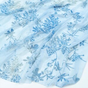 May include: A sheer, light blue scarf with a floral pattern in shades of blue and gray. The delicate fabric is draped, showcasing the intricate botanical design. The scarf's airy quality suggests a lightweight accessory.