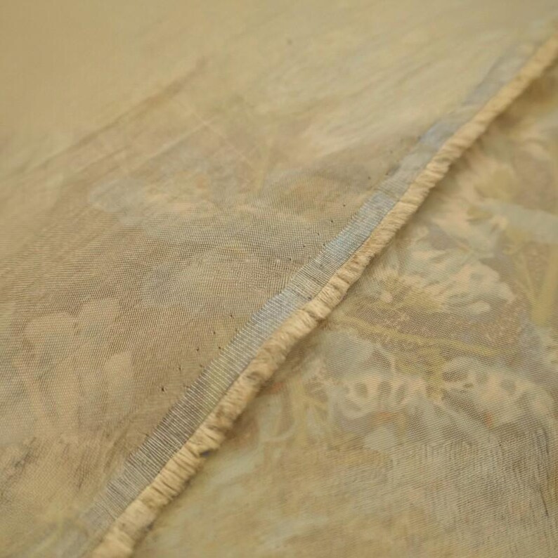 Puede incluir: Una tela ligera y transparente con un estampado floral en tonos beige y crema apagados. El material presenta un tejido visible y un borde acabado con un ribete azul claro, mostrando una textura delicada y semitransparente en todo el tejido.