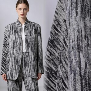 Könnte beinhalten: Eine langärmelige Jacke mit Stehkragen und einem abstrakten schwarz-weißen Pinselstrichmuster. Die Jacke verfügt über einen Vorderverschluss mit drei Knebelknöpfen und zwei Seitentaschen und wird über einem schlichten weißen Oberteil getragen.