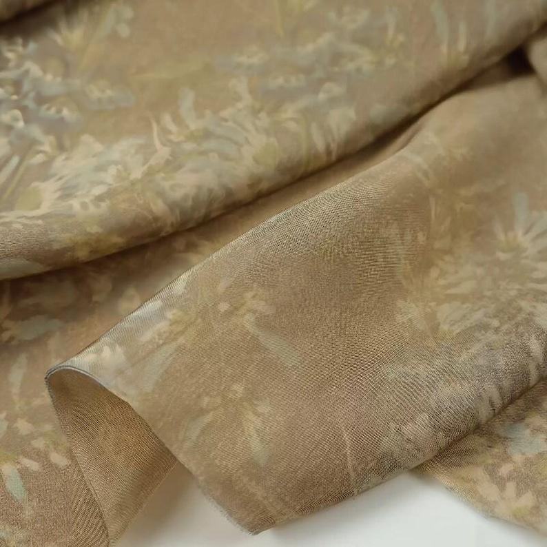 May include: Close-up of a folded fabric with a floral pattern. The fabric is a blend of tan, beige, and light green hues. The floral design appears to be a delicate, stylized botanical print. The material has a subtle sheen, suggesting a silk or satin weave.