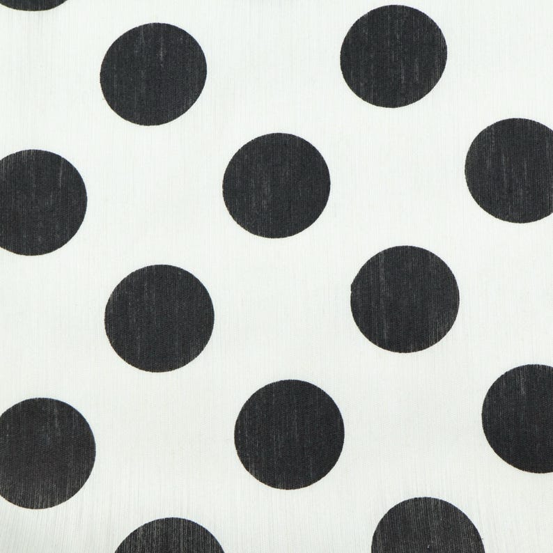 May include: Close-up of a white fabric with a repeating pattern of large, evenly spaced black polka dots. The dots are solid black and circular, creating a high-contrast design. The fabric appears to be lightweight.