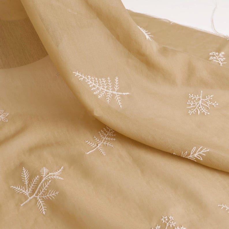 May include: Tan sheer fabric with white embroidered leaf patterns. The fabric is draped, showing the texture and translucence. The embroidery is detailed, creating a delicate and elegant design. Ideal for apparel or home decor.