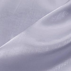 May include: Close-up of a light lavender fabric with a subtle sheen. The fabric is draped, creating soft folds and shadows. The material appears lightweight and slightly translucent, suggesting it could be used for clothing or decorative purposes.