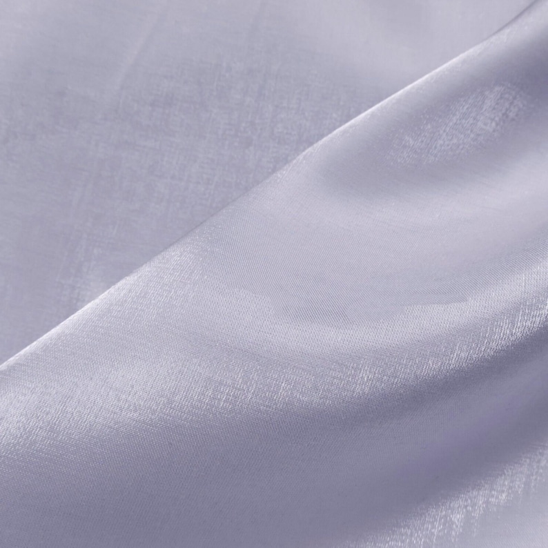 May include: Close-up of a light lavender fabric with a subtle sheen. The fabric is draped, creating soft folds and shadows. The material appears lightweight and slightly translucent, suggesting it could be used for clothing or decorative purposes.
