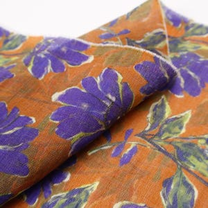 May include: Close-up of a folded fabric with a floral pattern. The fabric has a burnt orange background with large purple flowers and green leaves. The fabric appears to be lightweight and possibly silk or a similar material.