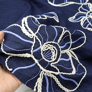 Può includere: Primo piano di tessuto blu navy con ricami floreali. Il ricamo presenta filo bianco e azzurro, creando un motivo decorativo strutturato. Il tessuto è piegato, mostrando i dettagli delle cuciture.
