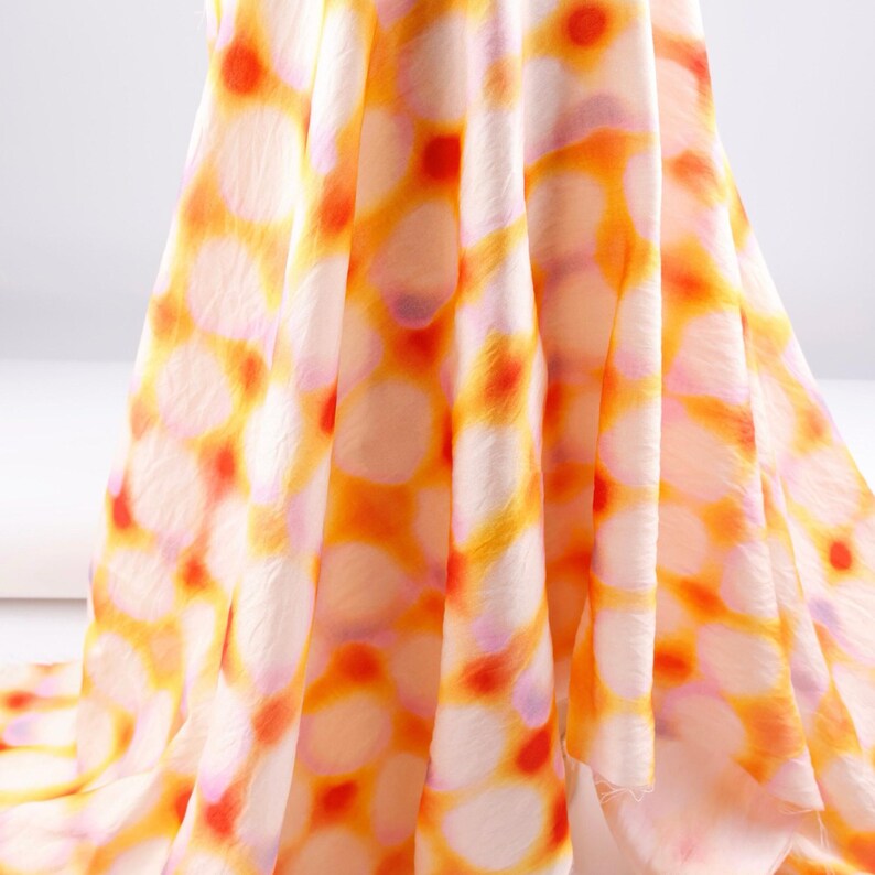 May include: A flowing fabric with a repeating pattern of orange and red circles on a white background. The fabric has a soft, delicate appearance, with visible folds and creases. The overall effect is light and airy.