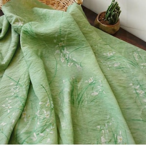 Peut inclure: Un tissu vert clair avec un motif floral d&eacute;licat. Le tissu pr&eacute;sente des fleurs blanches et des tiges et feuilles vertes, cr&eacute;ant un design naturel et a&eacute;r&eacute;. Le tissu est drap&eacute;, montrant sa texture et son flux.