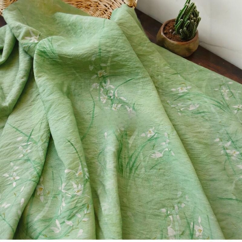 Peut inclure: Un tissu vert clair avec un motif floral d&eacute;licat. Le tissu pr&eacute;sente des fleurs blanches et des tiges et feuilles vertes, cr&eacute;ant un design naturel et a&eacute;r&eacute;. Le tissu est drap&eacute;, montrant sa texture et son flux.