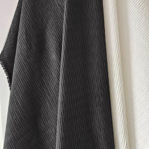 May include: Two pieces of fabric displayed side by side, one in solid black and one in solid white. Both fabrics feature a distinct, uniform, vertical pleated texture that creates a series of fine, parallel ridges across the entire surface.