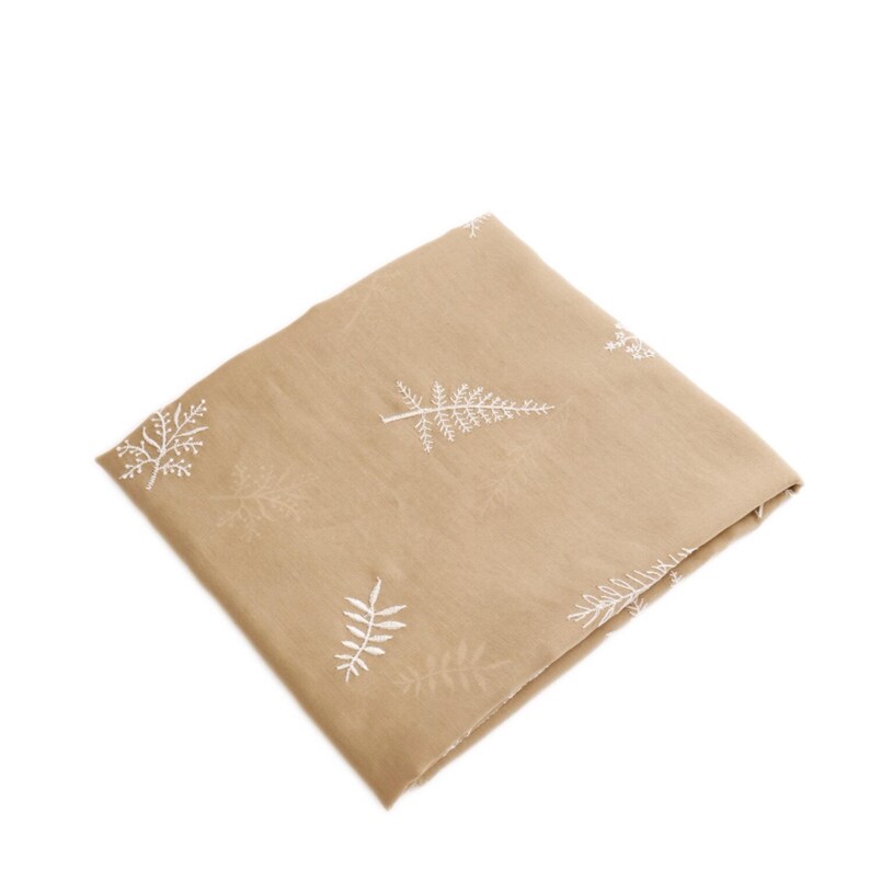May include: Folded tan fabric with a repeating pattern of white embroidered fern-like designs. The fabric is square and appears to be made of a soft material. The white embroidery provides a contrast against the tan background.