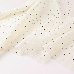 May include: Off-white fabric with a delicate, textured surface. The fabric is adorned with a pattern of small, evenly spaced black dots. The material is draped, creating soft folds and shadows, suggesting a lightweight textile.