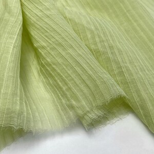 May include: Close-up of a light green fabric with a ribbed texture. The fabric is folded, showing the texture and the frayed edges. The color is a soft, pale green, and the fabric appears to be lightweight.