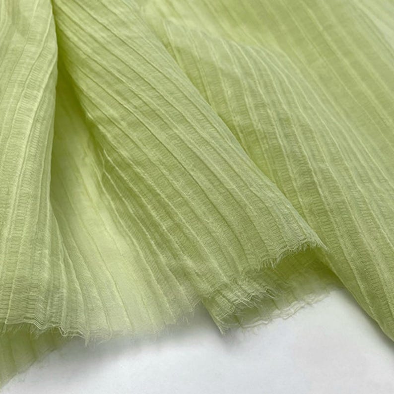May include: Close-up of a light green fabric with a ribbed texture. The fabric is folded, showing the texture and the frayed edges. The color is a soft, pale green, and the fabric appears to be lightweight.