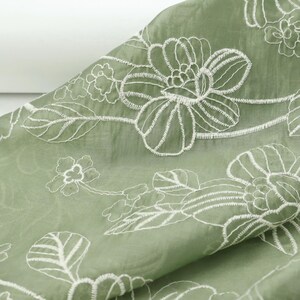 May include: A sage green fabric featuring a delicate white embroidered floral pattern. The design consists of large blossoms and leaves outlined in white thread, creating a continuous botanical motif across the smooth, slightly textured material.