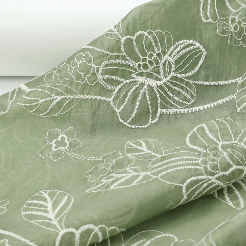May include: A sage green fabric featuring a delicate white embroidered floral pattern. The design consists of large blossoms and leaves outlined in white thread, creating a continuous botanical motif across the smooth, slightly textured material.