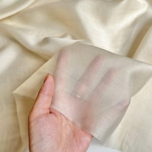 以下が含まれることがあります： 手元に、薄いクリーム色のシアー生地を持ったクローズアップ。生地は半透明で、指が部分的に透けて見えます。背景は同じ生地の大きなもので、柔らかい折り目があります。