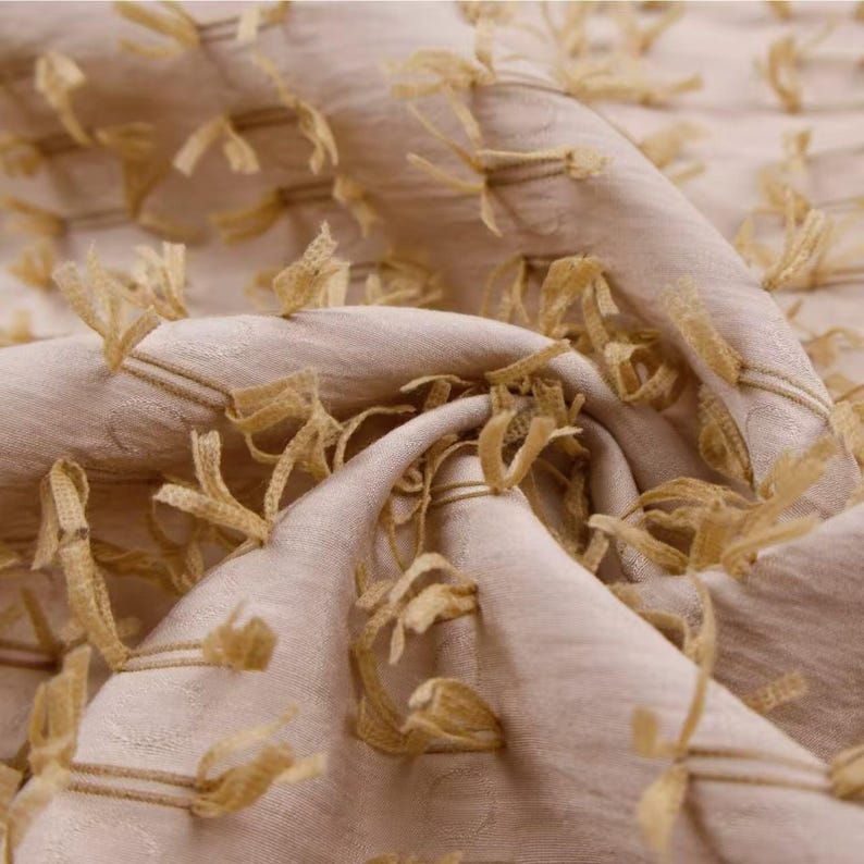 May include: A light beige fabric featuring a textured surface with numerous small, frayed, golden-yellow thread loops attached in a repeating pattern across the material, creating a dimensional and tactile appearance throughout the folded textile.