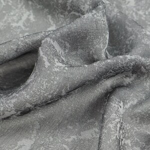May include: Close-up of a gray fabric with a textured surface and a subtle, abstract pattern in lighter shades of gray and white. The fabric is draped, showing folds and shadows, creating depth and visual interest.